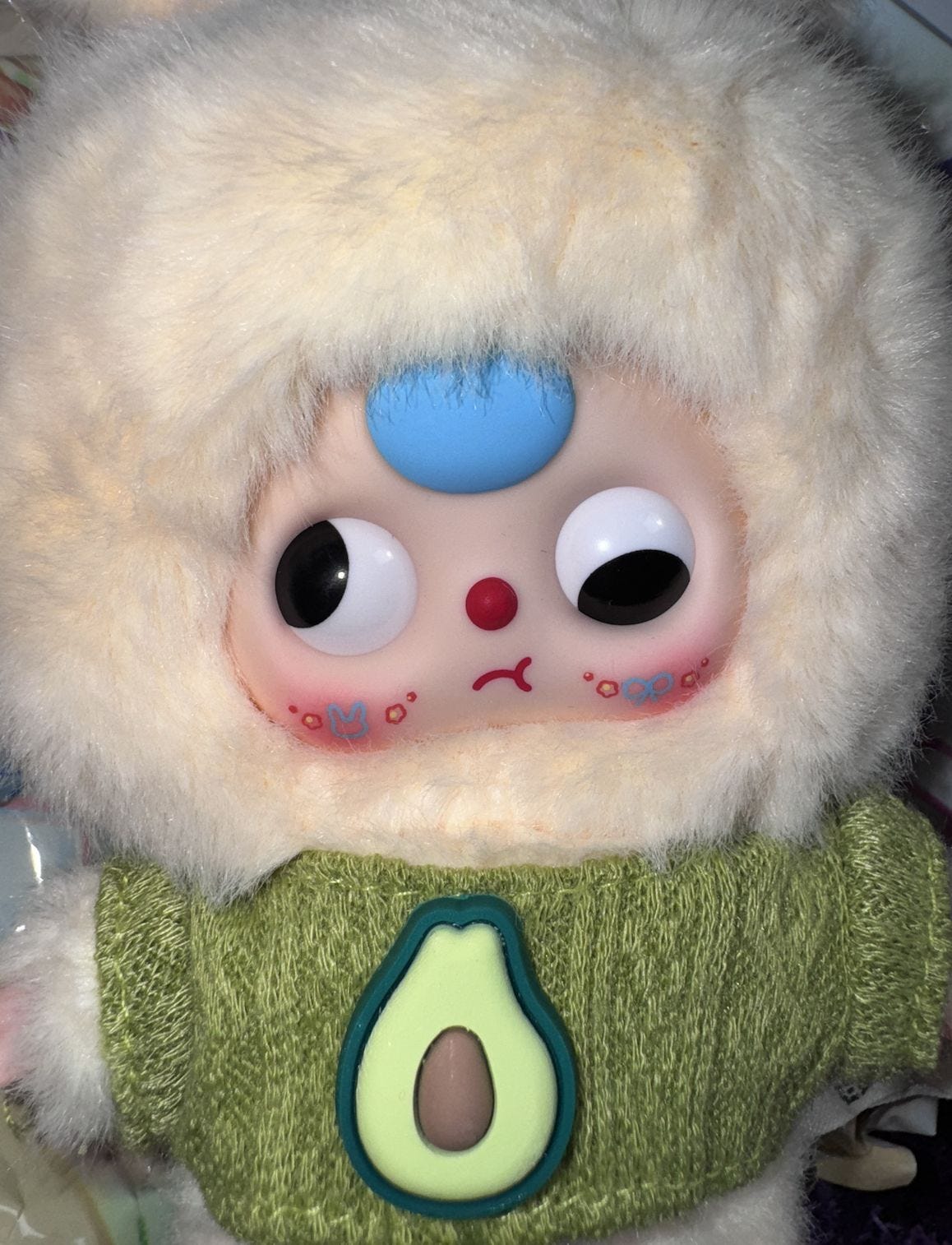Mid-framed shot of a Baby Three plush doll: cream-colored furry hood, blue circle on the forehead, wide black-pupil eyes, tiny red nose and mouth, wearing a knitted green sweater with a stylized avocado patch. 