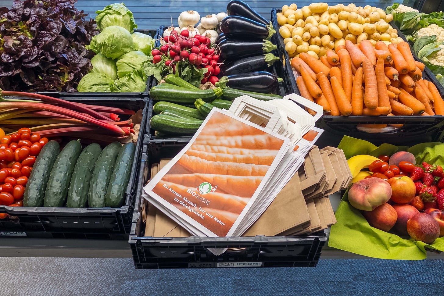 Bunt bestückter Marktstand mit frischem Bio-Gemüse und Bio-Obst in schwarzen Kisten: zu sehen sind unter anderem Blattsalate, Eisbergsalat, Radieschen, Auberginen, Kartoffeln, Karotten, Zucchetti, Rhabarber, Gurken und Cherrytomaten. In der Mitte liegt eine Papiertüte von Bio Suisse mit dem Knospe-Logo.