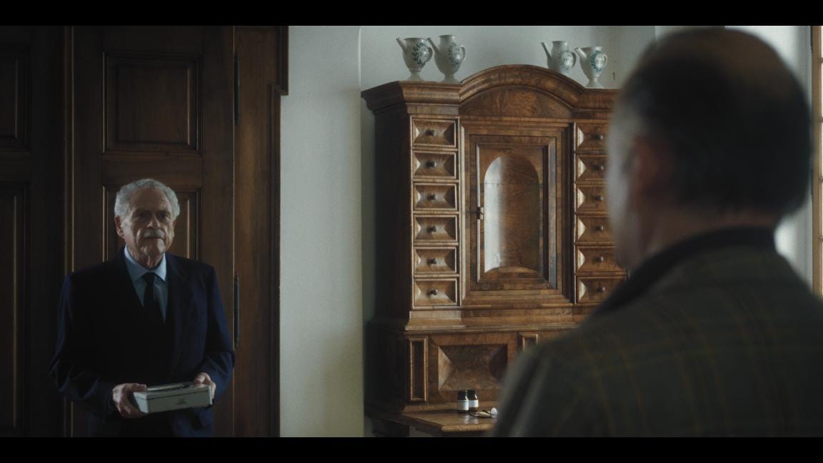 An elegant old man walking towards a middle-aged man with a cookie tin in his hand. 