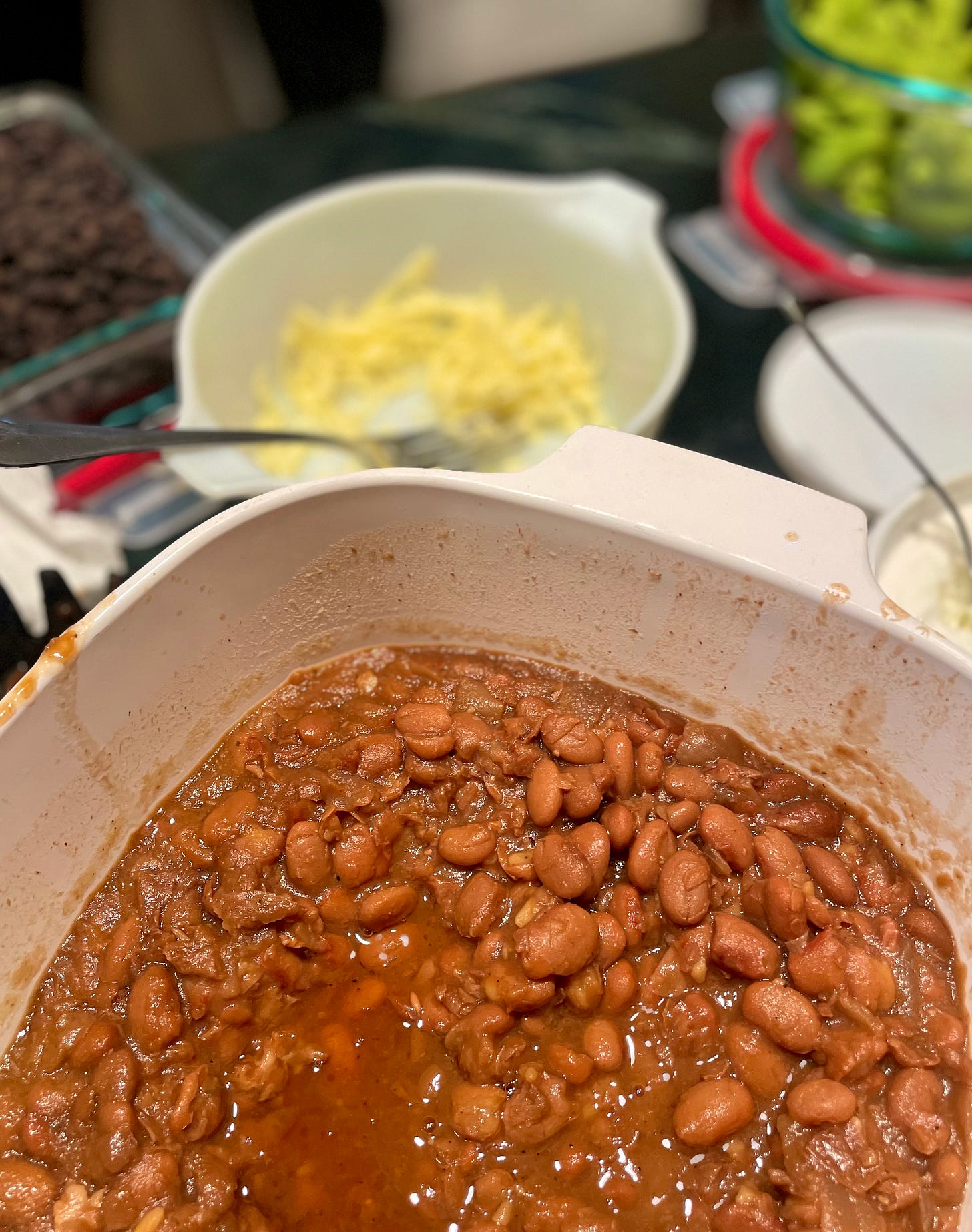 Close up of author's finished beans on counter top at potluck. Blurry background images include bowl of shredded cheese, bowl of black beans and bowl of lettuce