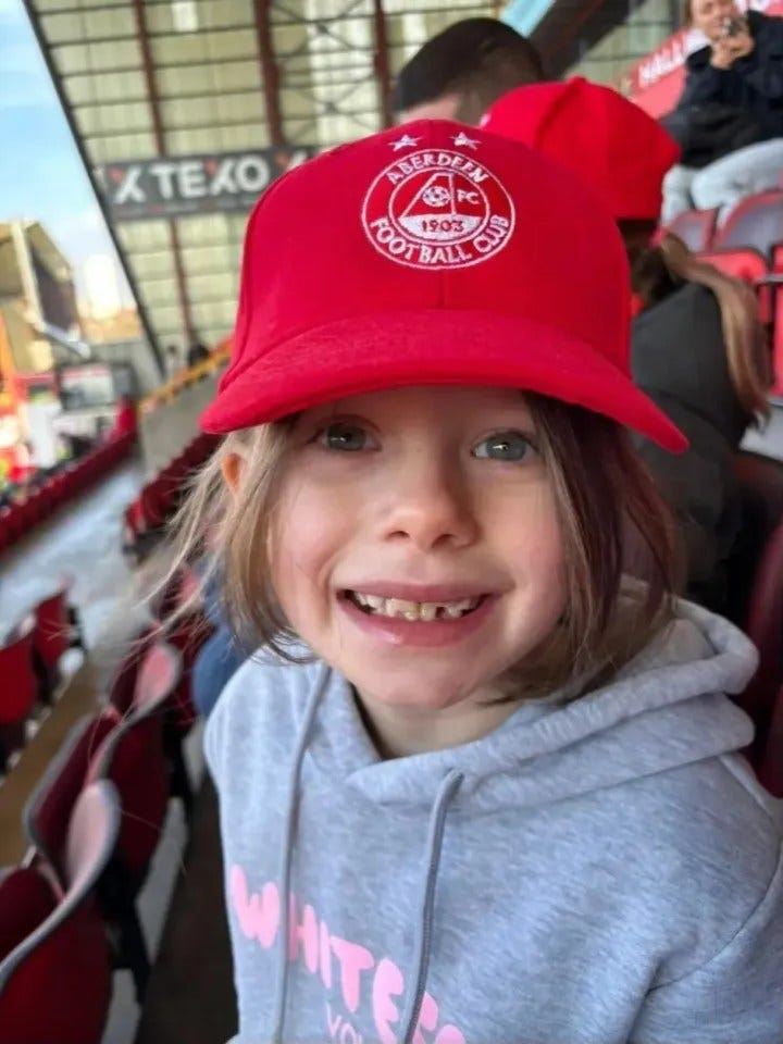 A young girl wearing an Aberdeen Football Club hat smiles. A young girl wearing an Aberdeen Football Club hat smiles.