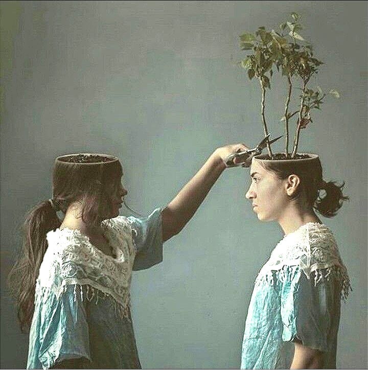 Surreal image of two women in matching dresses. One has an open head filled with soil and a growing plant. The other, whose own head is hollow and empty, trims the plant growing from the first woman's head, as if harvesting her thoughts.