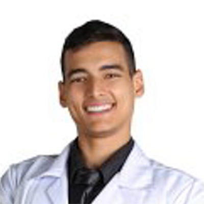 Homem jovem, de cabelos pretos e curtos, sorrindo e olhando para a câmera. Ele veste um jaleco branco sobre uma camisa escura e está com os braços cruzados. Homem jovem, de cabelos pretos e curtos, sorrindo e olhando para a câmera. Ele veste um jaleco branco sobre uma camisa escura e está com os braços cruzados.