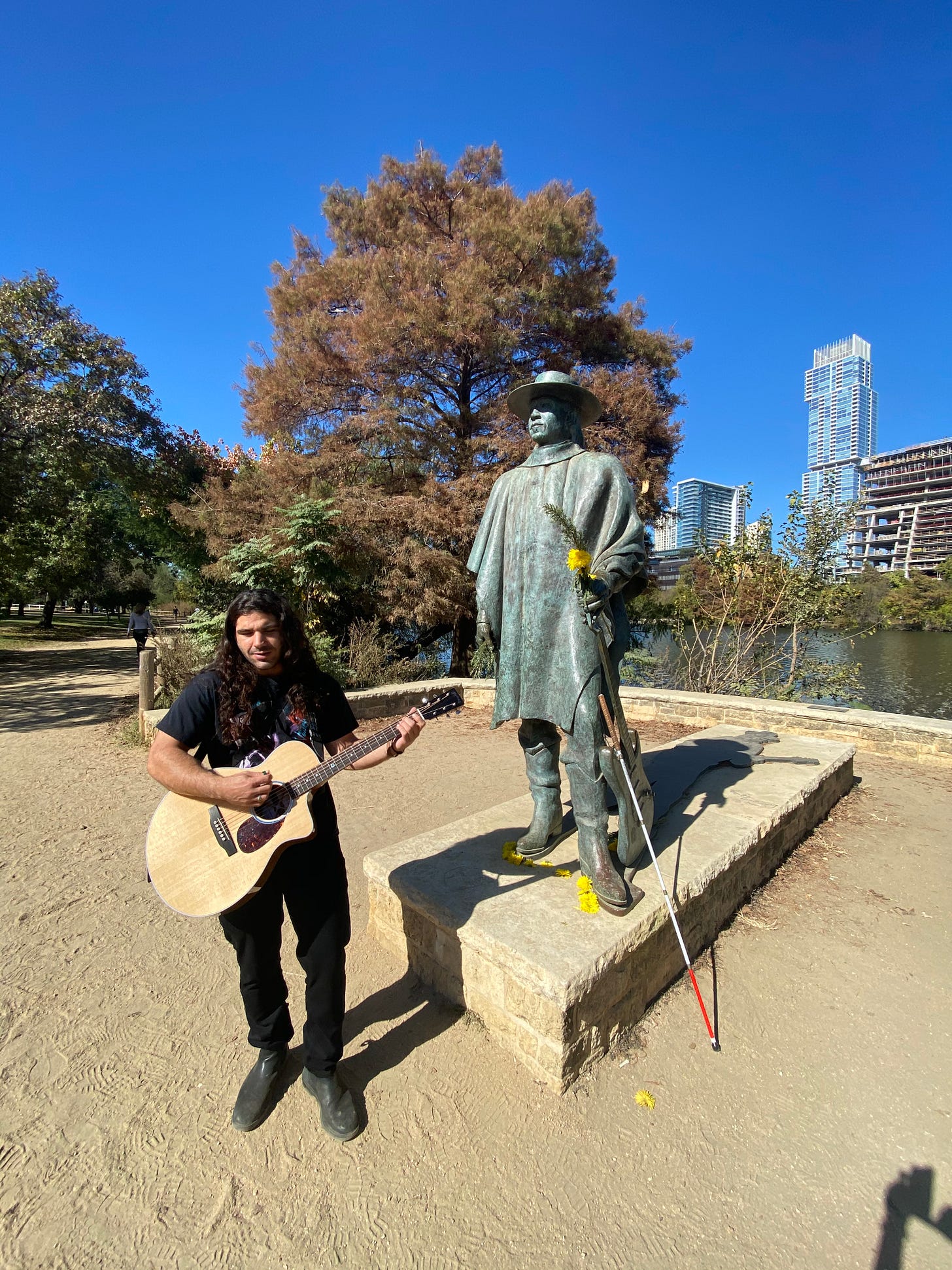 anthony standing in front of the stevie ray vaughn status with yellow flowers on stevies hands -- ant is holding his guitar and smiling up at his hero
