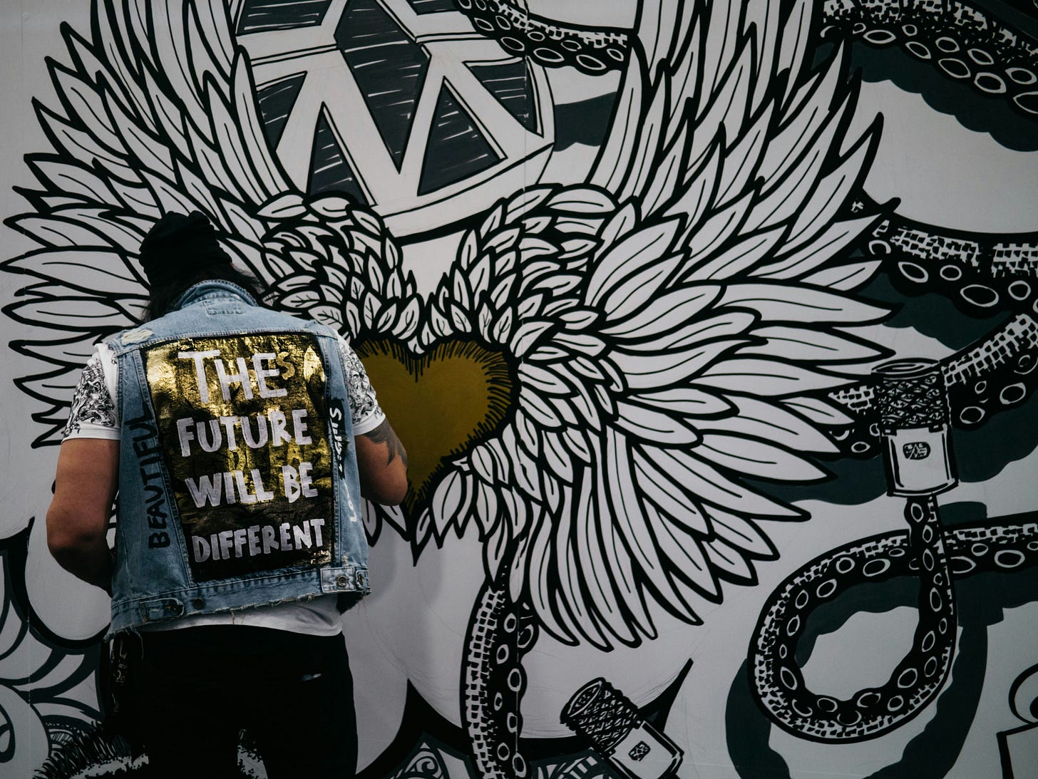 A man with his back to the camera works on a large black, white and gold art installation featuring, at its centre, a winged heart. The man wears a denim vest which reads "the future will be different". A man with his back to the camera works on a large black, white and gold art installation featuring, at its centre, a winged heart. The man wears a denim vest which reads "the future will be different".