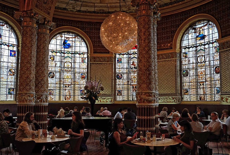 People drink tea and chat at tables in a grand museum cafe, with ornate carved pillars and a spherical pendant lamp. People drink tea and chat at tables in a grand museum cafe, with ornate carved pillars and a spherical pendant lamp.