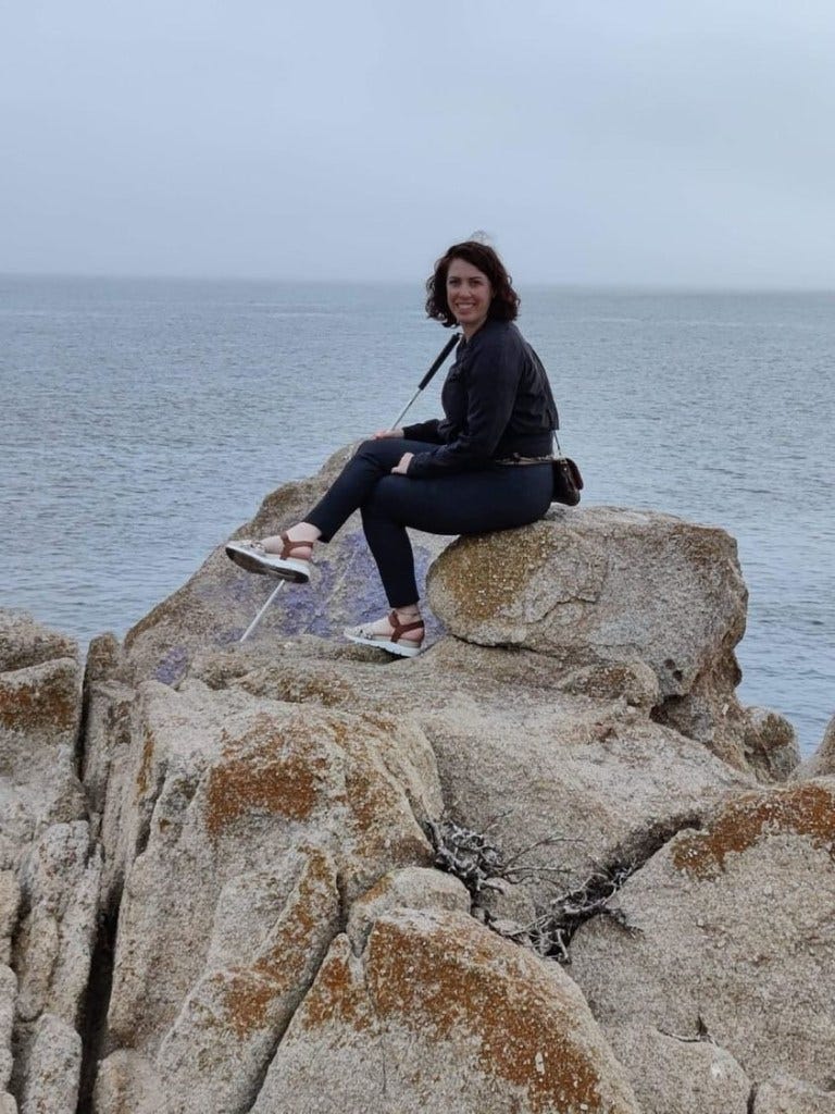 Becky, a white woman with curly, brown, shoulder-length brown hair sits on large rocks at the sea. She is dressed in black and wearing sandals. Her white cane rests in her right hand. Becky, a white woman with curly, brown, shoulder-length brown hair sits on large rocks at the sea. She is dressed in black and wearing sandals. Her white cane rests in her right hand.