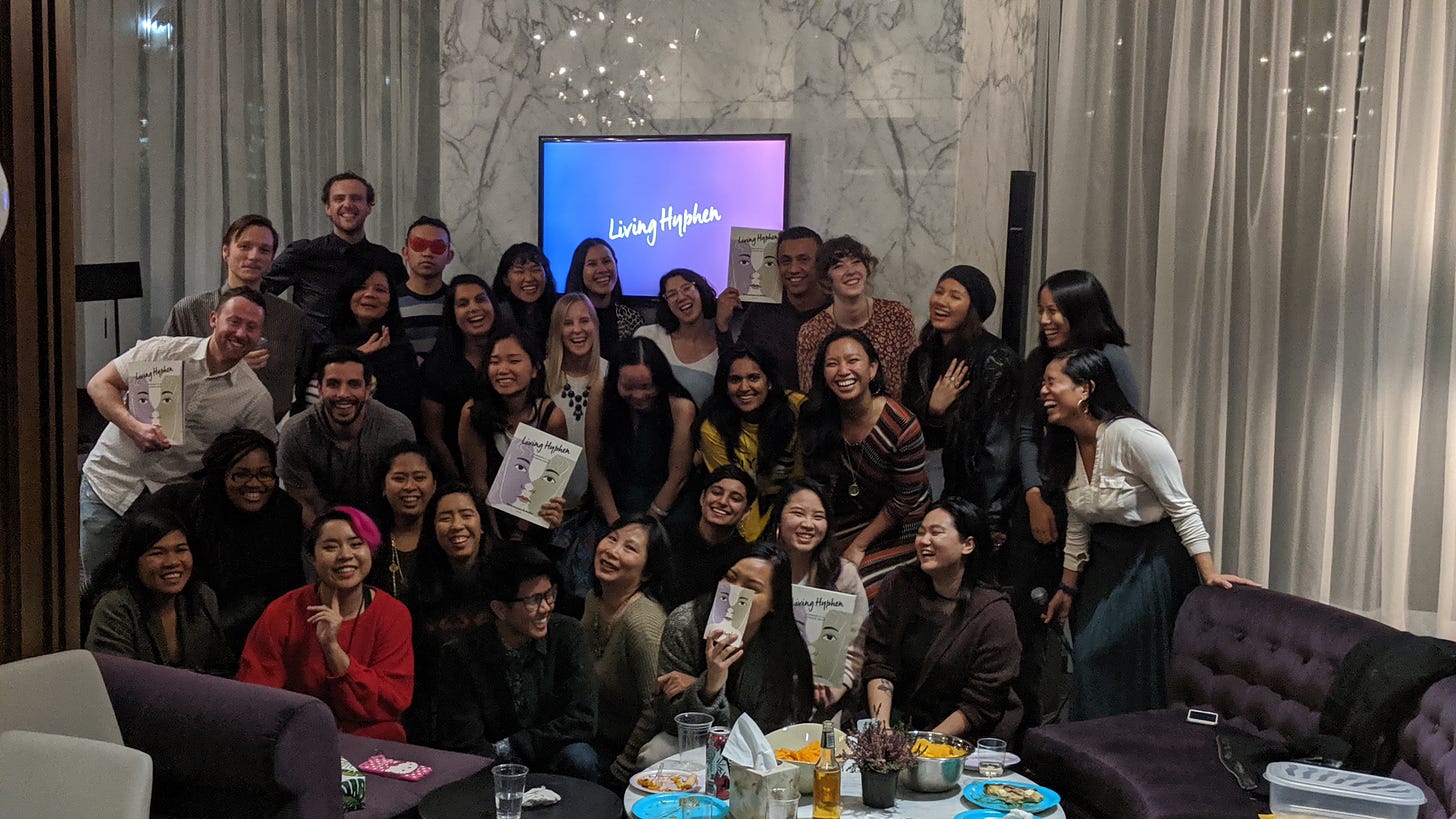 Living Hyphen community standing in a big group laughing and holding up copies of the magazine. Living Hyphen logo is on a screen in the background.