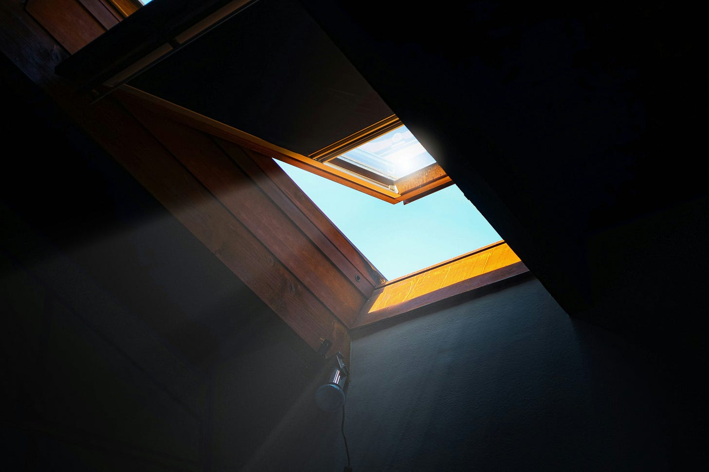 A skylight is open in a dark room, letting sunlight stream in. The frame is wooden, and a soft beam of light cuts across the shadows toward the floor, suggesting hope and illumination.