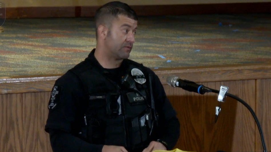 Seattle Police officer Anthony Ducre, buzz cut, 40s, in uniform, stands at a lectern in an auditorium