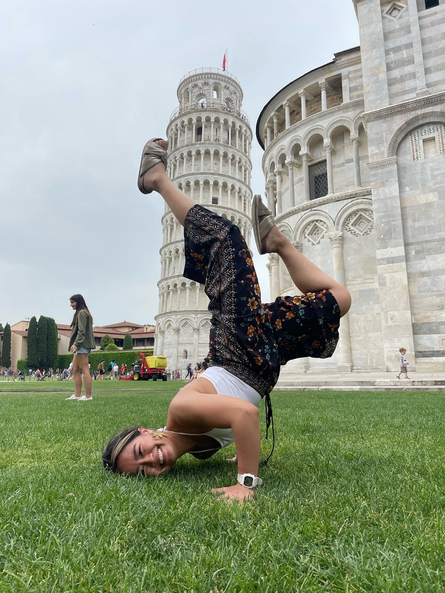 Joelle doing a freeze, a breakdancing pose where she balances on the side of her temple and her hands and has her legs in the air, with the Leaning Tower of Pisa between her feet