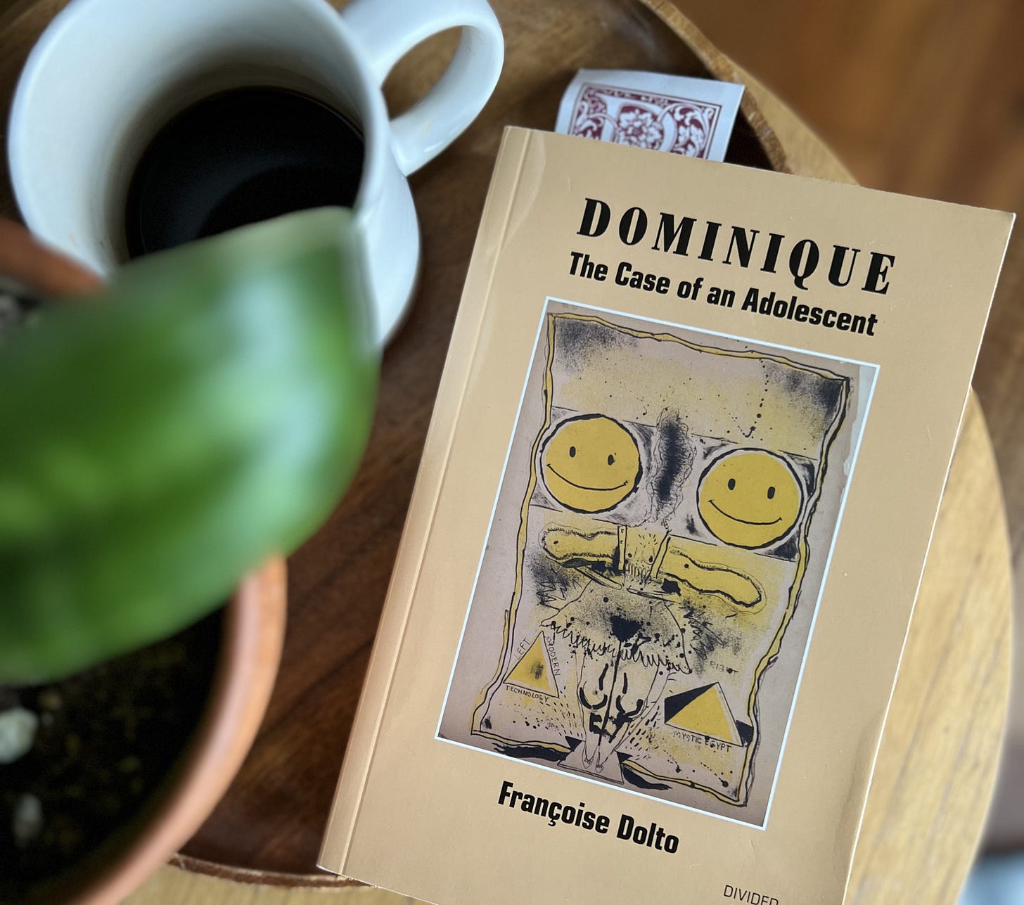 The book Dominique by Francoise Dalto sits beside a cup of coffee and a blurry houseplant