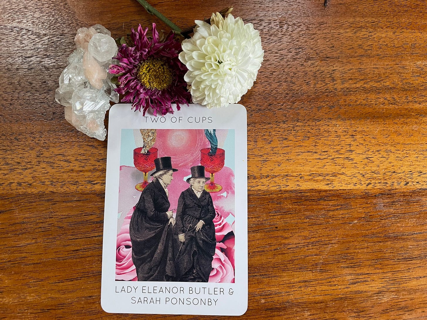 A purple flower, a white flower, and a pink and clear quartz crystal sit atop a Two of Cups tarot card with an old-timey rendering of two women dressed in top hats and tuxedo dresses. Lady Eleanor Butler & Sarah Ponsonby