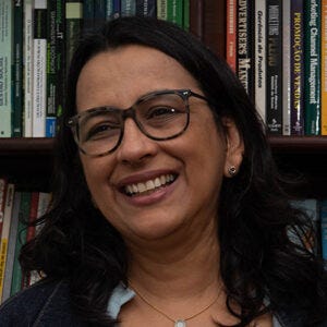 Mulher de pele clara e cabelos escuros, levemente ondulados. Usa óculos de armação escura e esboça um leve sorriso. Fundo, uma estante de livros.