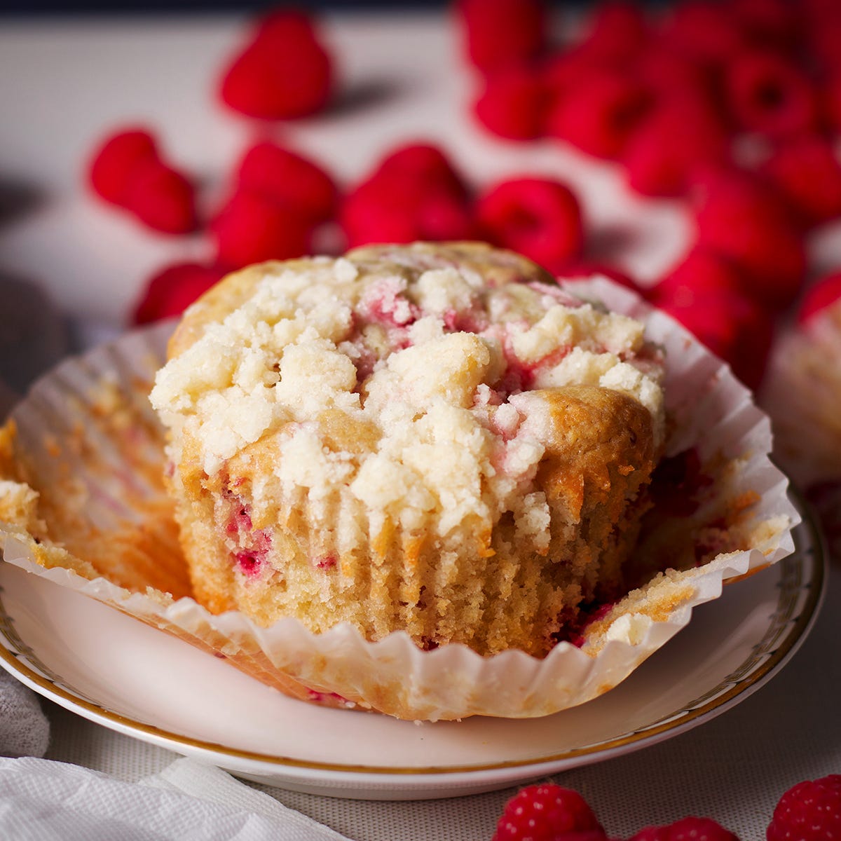 A freshly baked raspberry muffin on a plate next to a pile of fresh raspberries.