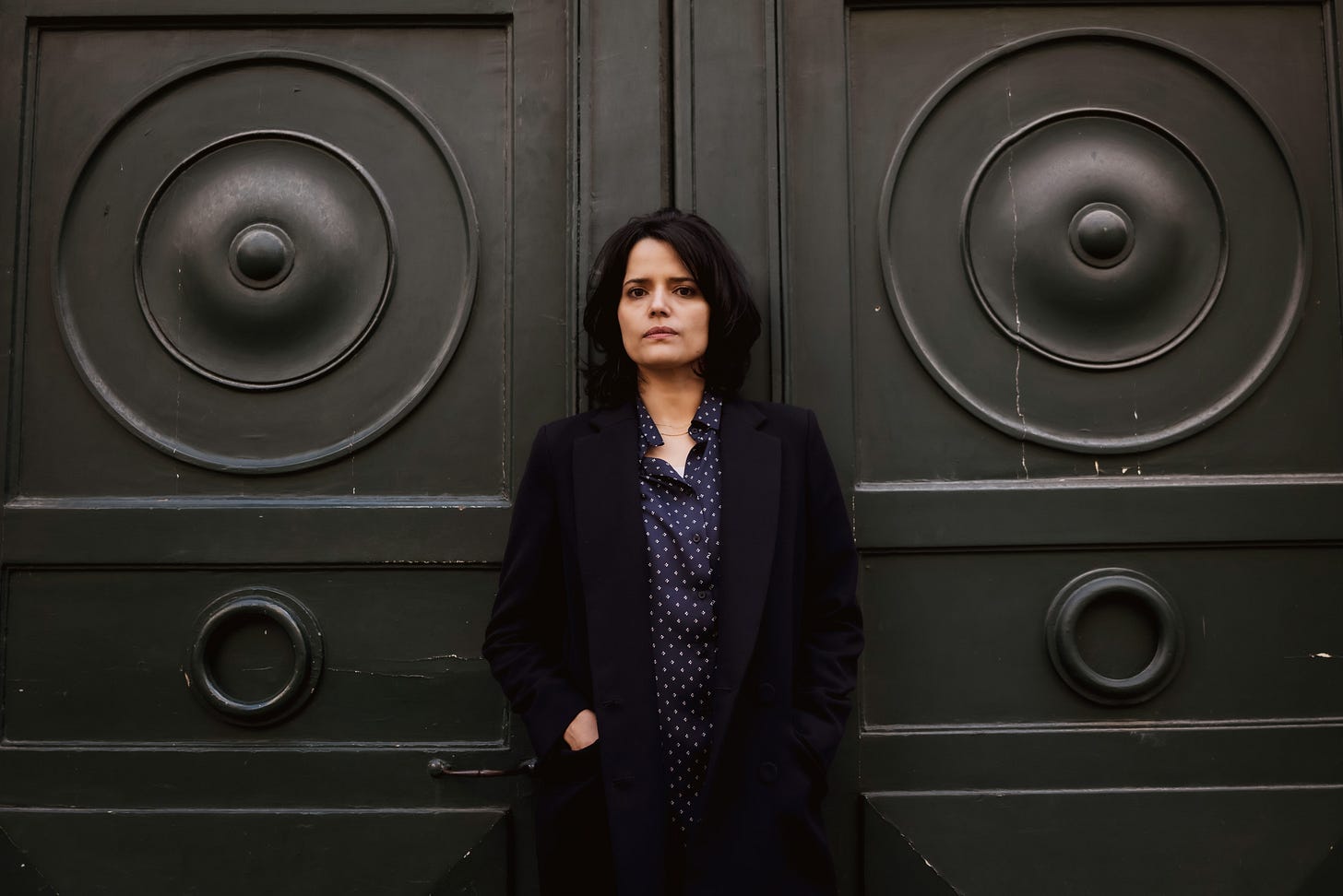 Portrait de Haïfa Tlili en Mars 2024. Plan américain. Elle se trouve à l'extérieur contre une grande porte noire. Elle est vêtue d'un manteau noire et d'une chemise bleue à pois blanc. Elle a les cheveux noirs mi-longs.