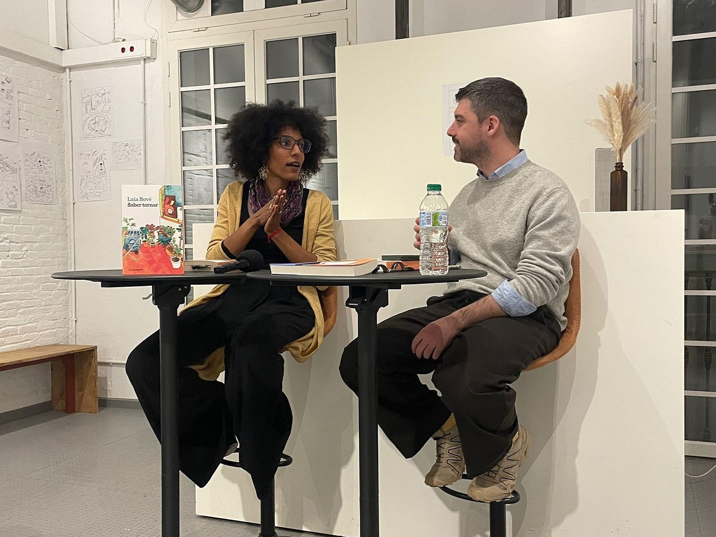 aia Bové and David Moragas speaking together at a book presentation event, both seated and smiling, in a warmly lit bookstore setting in Barcelona. 
