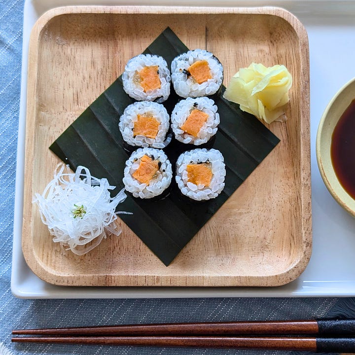 Two types of hosomaki sushi rolls. One is filled with roasted butternut squash and candied ginger; the other is a classic cucumber roll topped with sesame seeds. Both are sliced into bite-sized pieces and arranged on a wood plate.