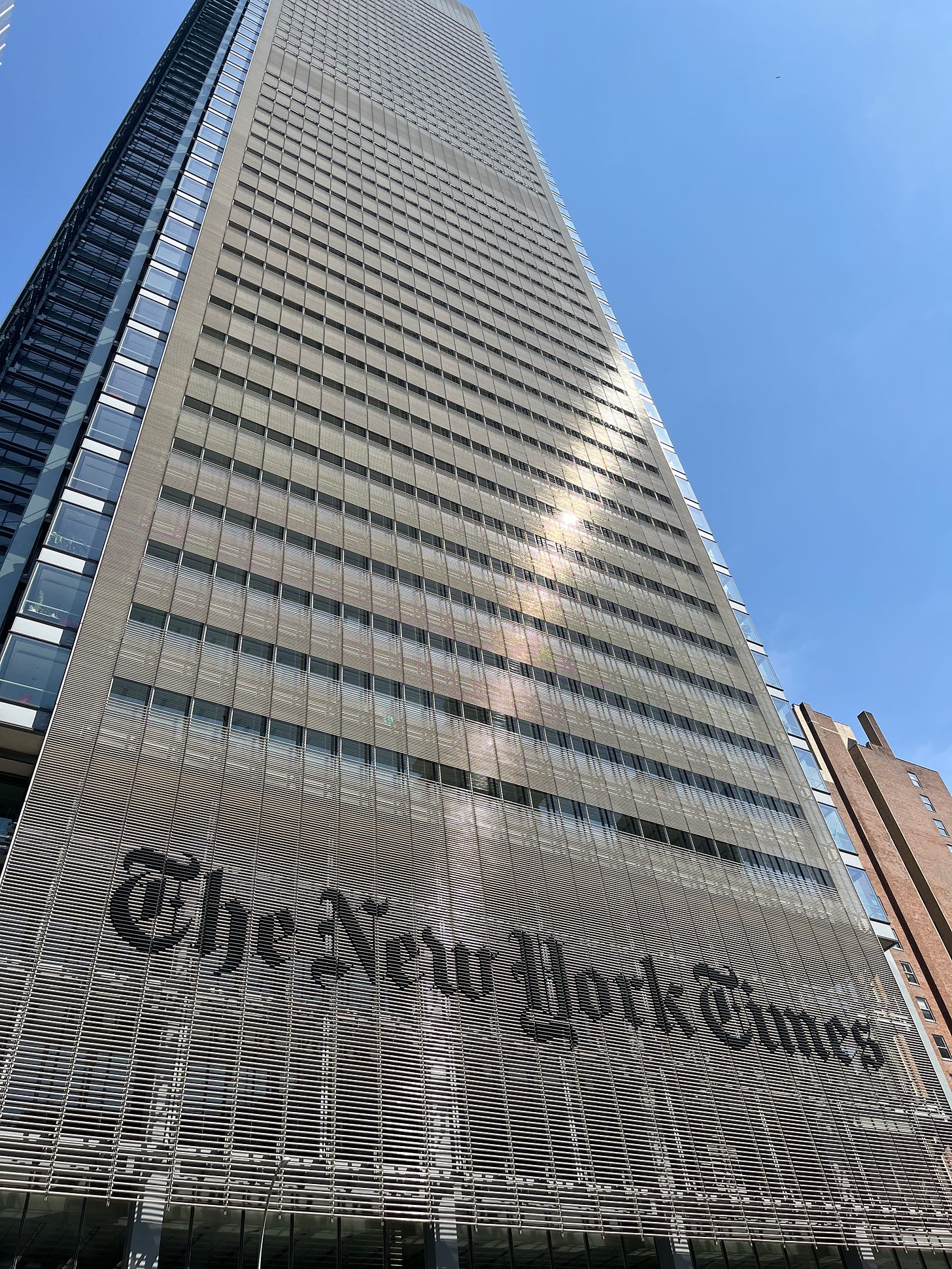 Upward view of The New York Times building in Manhattan, showing its tall glass and metal facade with the newspaper’s logo prominently displayed on the lower exterior. Upward view of The New York Times building in Manhattan, showing its tall glass and metal facade with the newspaper’s logo prominently displayed on the lower exterior.