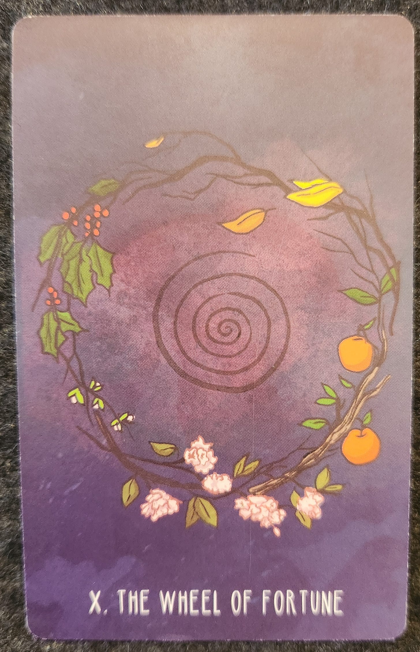 A tarot card labelled X The Wheel of Fortune. Branches form a circle, with each section of the branches growing different plants. There are green leaves and red berries. Small yellow buds. Small green leaves with fully bloomed pink flowers. Small green leaves with two plump oranges, and a section with the leaves are yellowing and falling off. In the centre of the circle is a black spiral. 