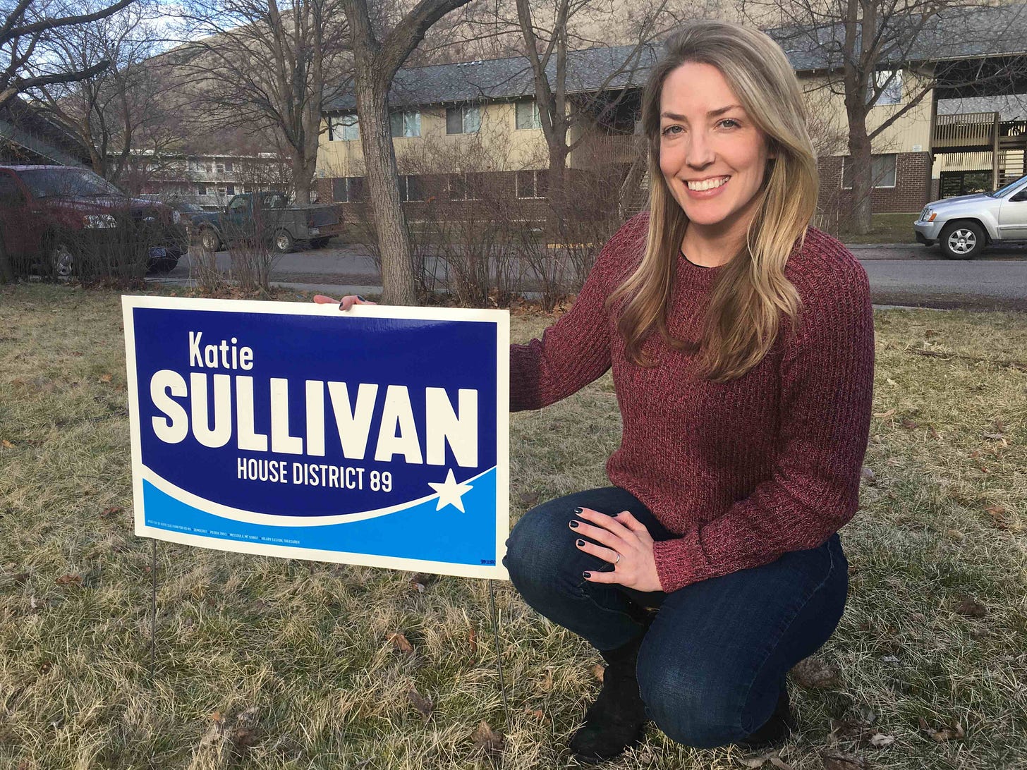woman with blonde hair in red sweater smiling next to a yard sign bearing her name