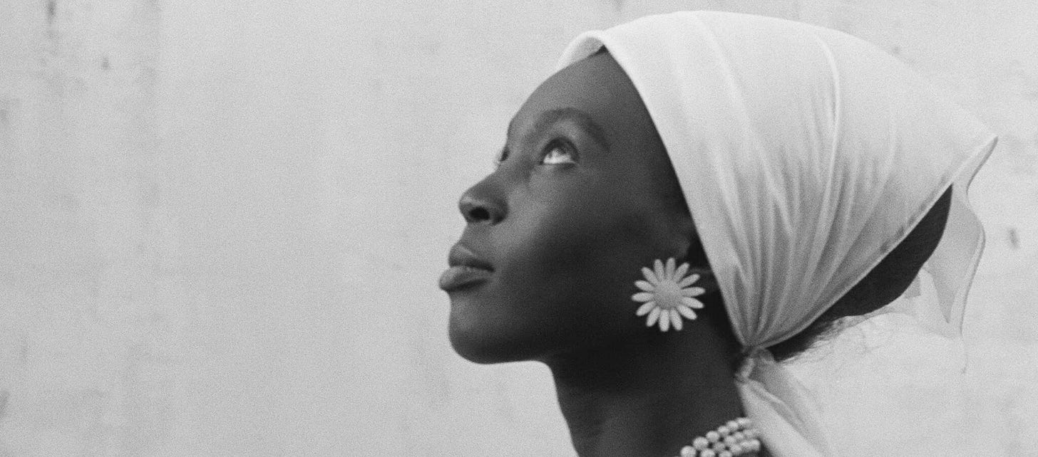 A black-and-white still of the profile of a young Black woman, who wears a head scarf, flower earrings, and a pearl necklace.