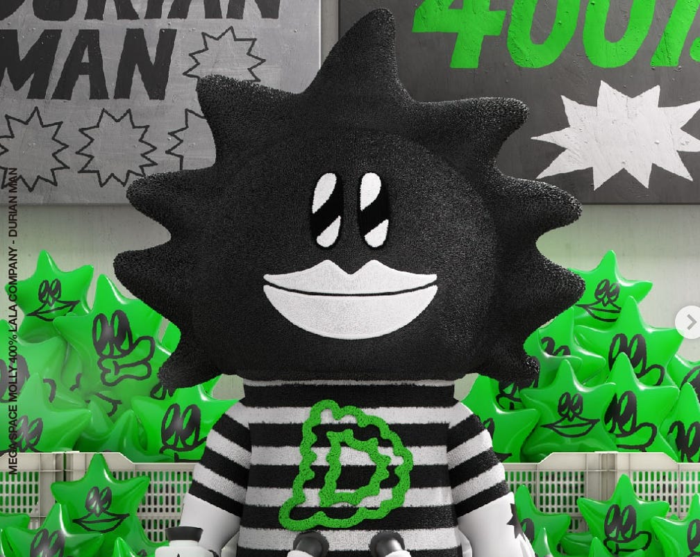 A 400% Mega Space Molly “Durian Man” figure from LALA Company stands in front of a wall decorated with comic-style signs and bright green star-shaped balloons. The figure features a spiky black head, a wide cartoonish smile, and wears a black-and-white striped shirt with a green “D” logo on the chest. A 400% Mega Space Molly “Durian Man” figure from LALA Company stands in front of a wall decorated with comic-style signs and bright green star-shaped balloons. The figure features a spiky black head, a wide cartoonish smile, and wears a black-and-white striped shirt with a green “D” logo on the chest.