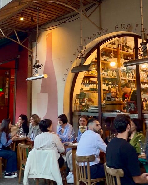 Photos inside In Vino wine bar in Yerevan, including a hand holding a bottle of Trinity Canyon Areni Noir, staff pouring wine behind shelves stocked with Armenian and international bottles, friends toasting with glasses of white wine, a long tasting table set with glassware beneath a sign reading 'The Renaissance of Armenian Wine Making,' and a wall of photos honoring winemakers