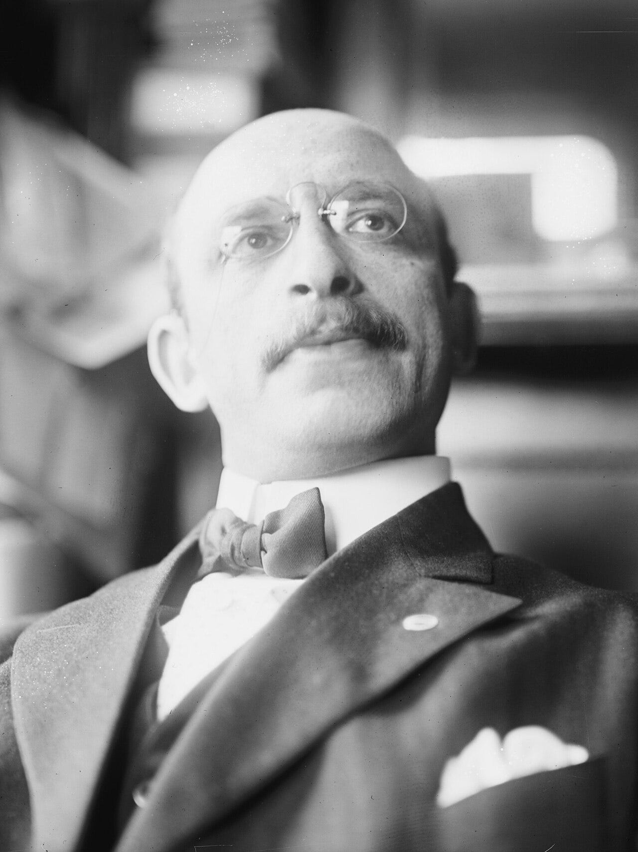 A man in his 40s with balding hair and a thick mustache, wearing circular glasses and a suit with a bowtie and pin, looks upward past the camera.