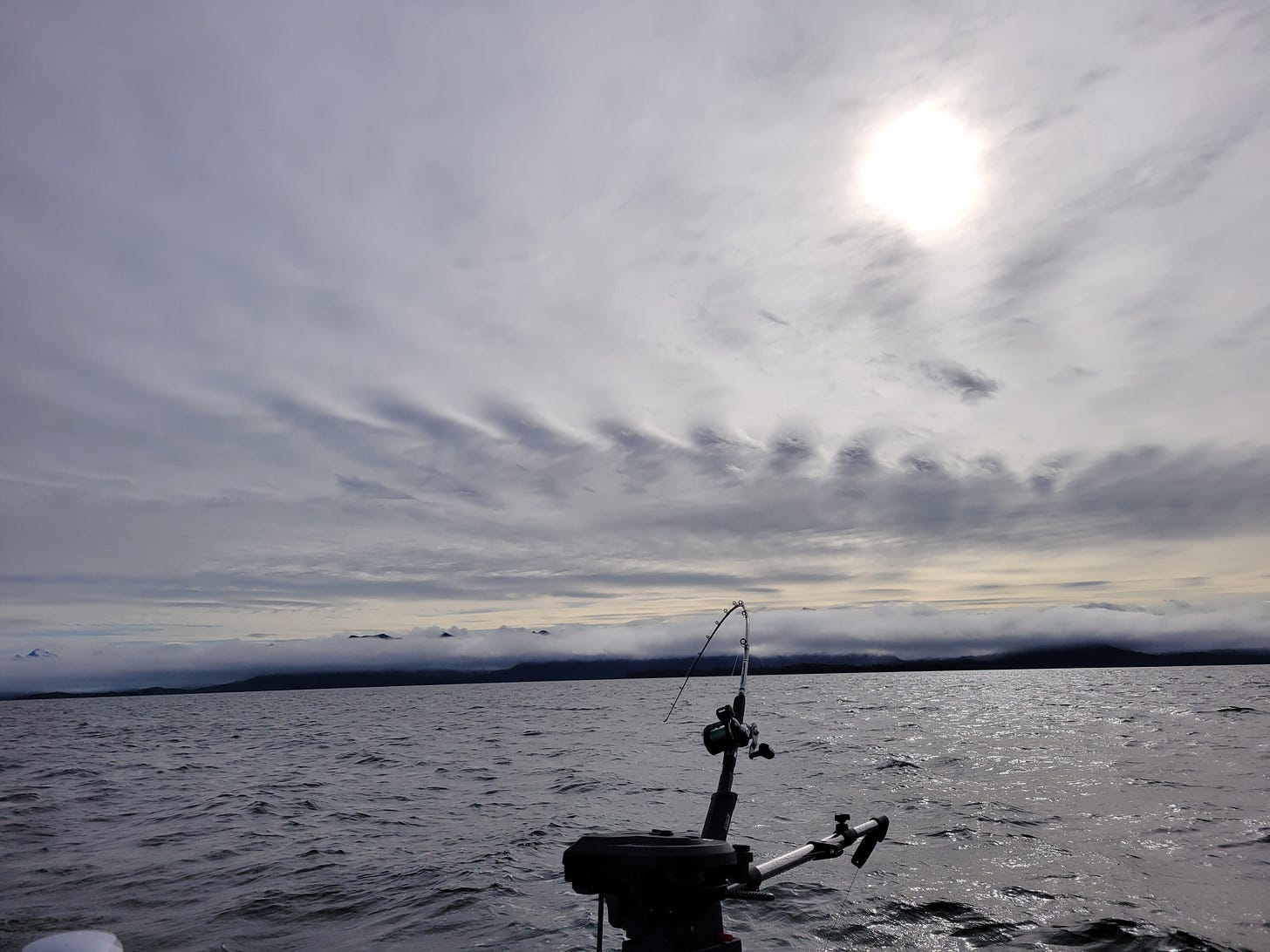 lines of clouds obscure a hazy sun sitting over a stretch of calm ocean. In the foreground, a fishing pole bends towards the water from it's stand on an electric downrigger. A great distance away low hanging clouds block most of the view of a coastal mountain range