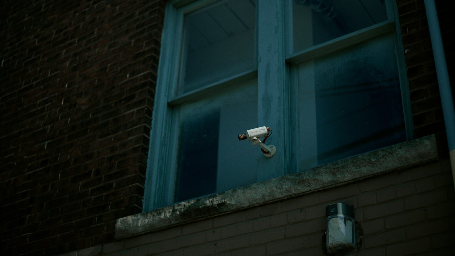Photo of a single surveillance camera mounted on a window frame near a brick wall.