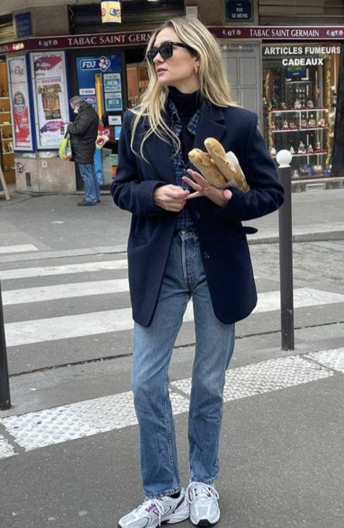 Influencer Anne-Laure Mais crosses a Parisian street wearing a navy blazer, straight-leg jeans, grey sneakers, and sunglasses, holding two baguettes in hand. Influencer Anne-Laure Mais crosses a Parisian street wearing a navy blazer, straight-leg jeans, grey sneakers, and sunglasses, holding two baguettes in hand.