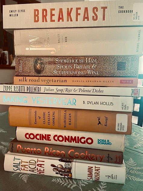 The cover of the first edition of Joy of Cooking, showing a stylized image of St. Martha of Bethany; copies of antique-looking cookbooks, and a stack of cookbooks on a dining room table