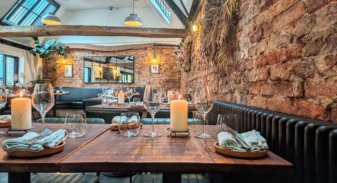 A restaurant with wooden tables with glasses, cutlery, and candles on it, and a tall ceiling with wooden beams