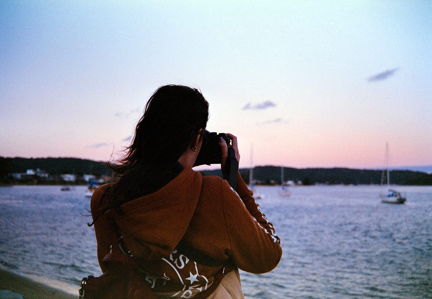 Josie taking photos of the last light of blue hour with her original Ricoh KR-10M. She’s facing towards the horizon wearing a brown hoodie, with the camera raised to her face. The sky is brilliant shades of blue and peach. Josie taking photos of the last light of blue hour with her original Ricoh KR-10M. She’s facing towards the horizon wearing a brown hoodie, with the camera raised to her face. The sky is brilliant shades of blue and peach.
