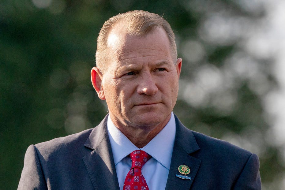 Headshot of Rep. Troy Nehls in a dark suit and red tie with a congressional lapel pin, trees blurred behind.