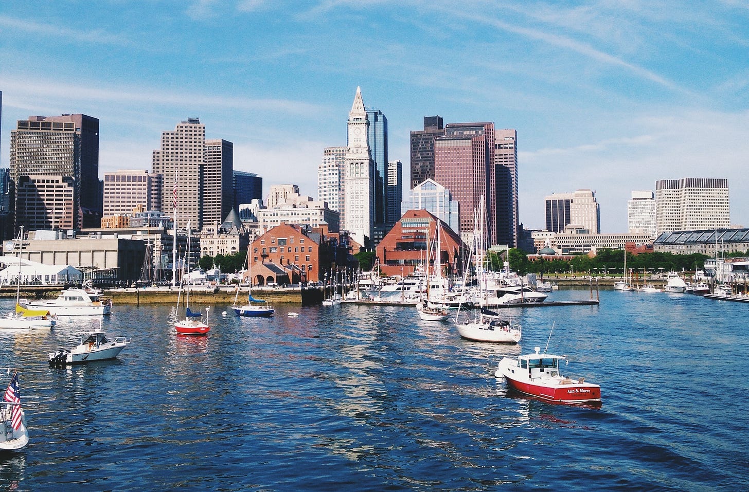 Ancored sail boats in a harbor with the city skyline in the background.