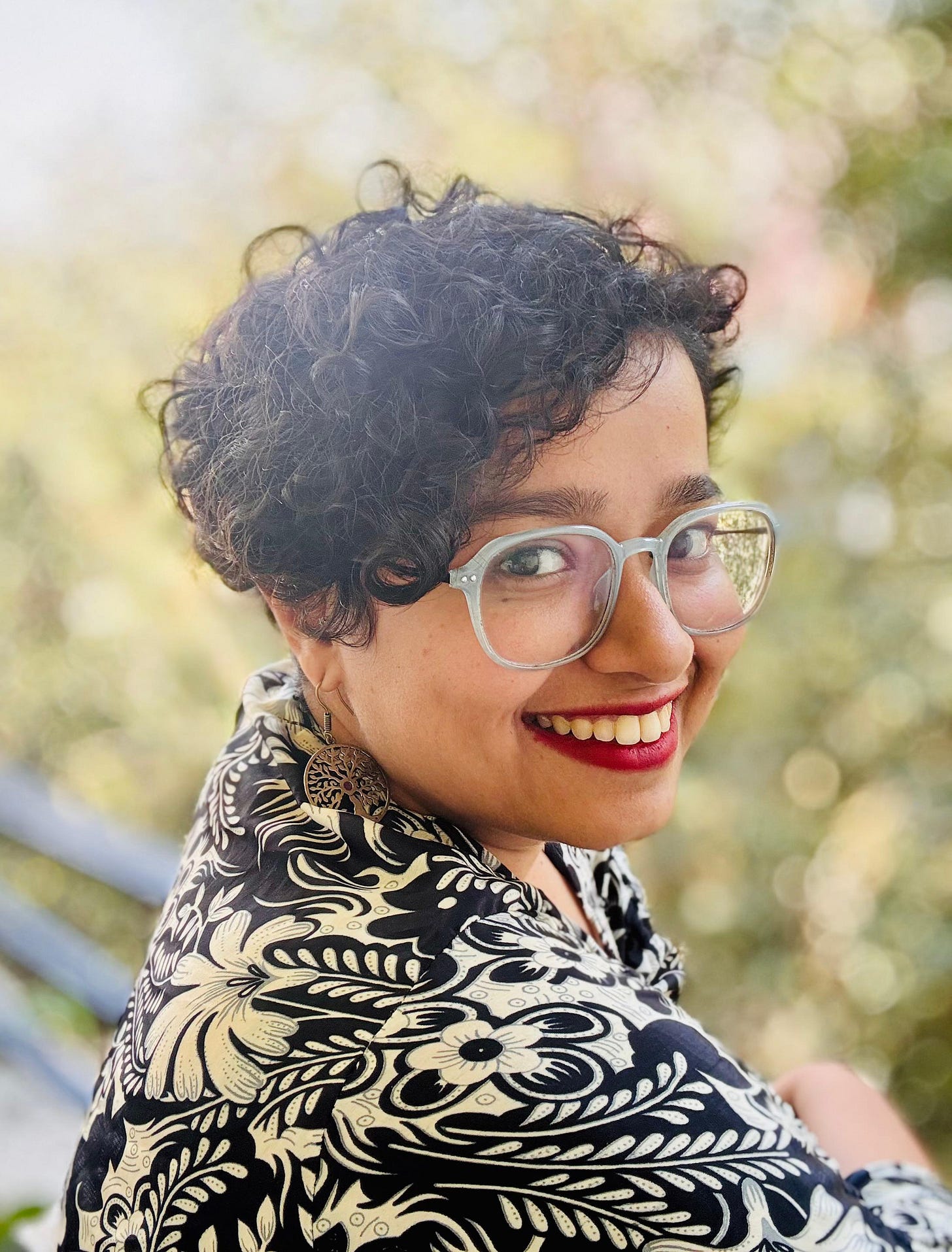 Raju, a young Indian woman with short curly hair and grey rimmed glasses is dressed in a floral black and white printed top and smiling into the camera.