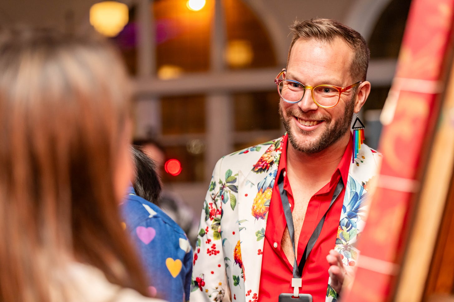 A white perseon with short hair and a beard is smiling, mid conversation with a blurred figure in the foreground. They are wearing rainbow glasses, a suit blazer that is white with fruit and flowers on it and a bright red shirt.