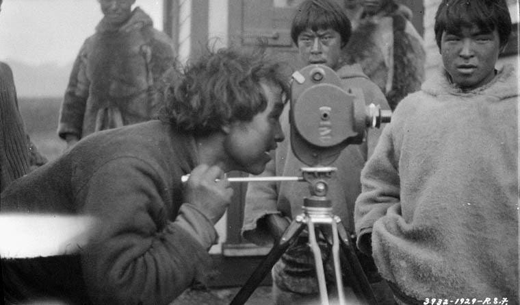 Man in foreground looking through camera on tripod, in front of several younger men in warm clothing