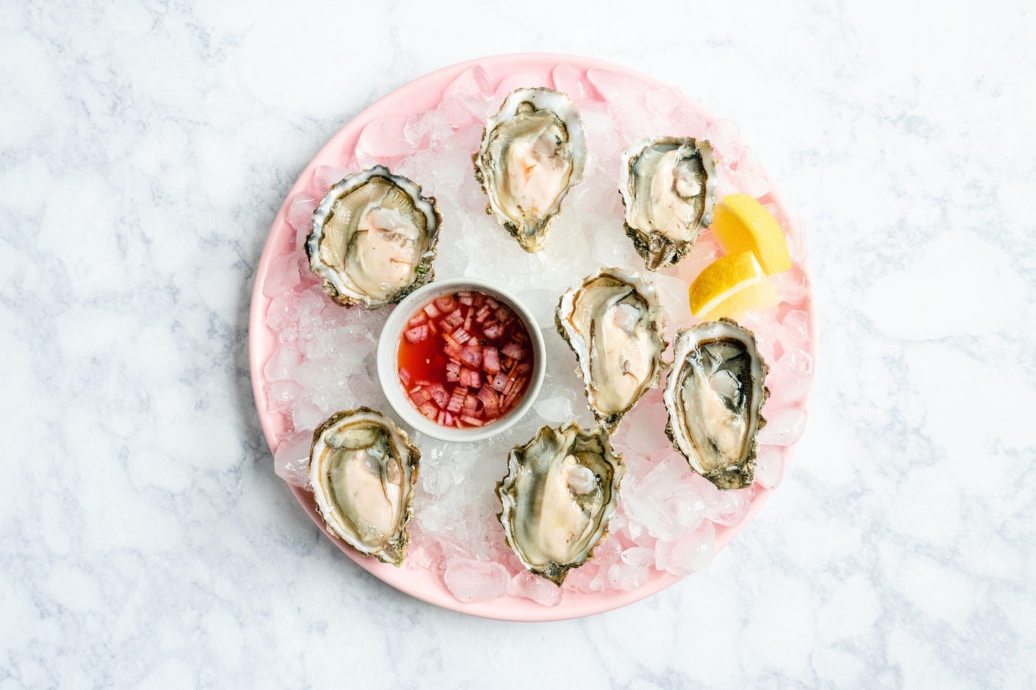 Pink plate with 7 oysters on the half shell, a wedge of lemon and a small dish of sheer red vinaigrette.