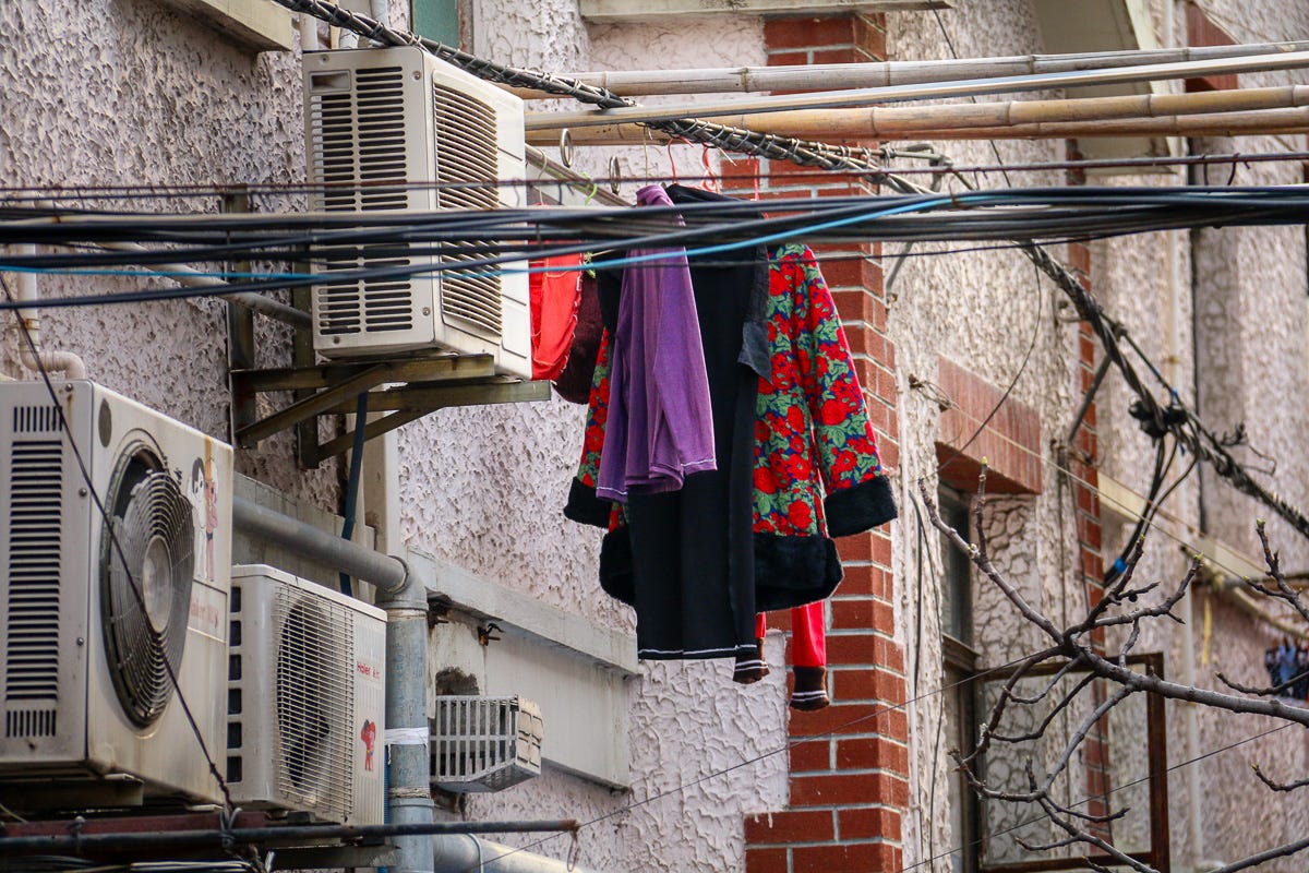 A striking black and red floral coat hangs from a pole beneath tangled power lines and A/C units. Fashion meets function above the sidewalk.
