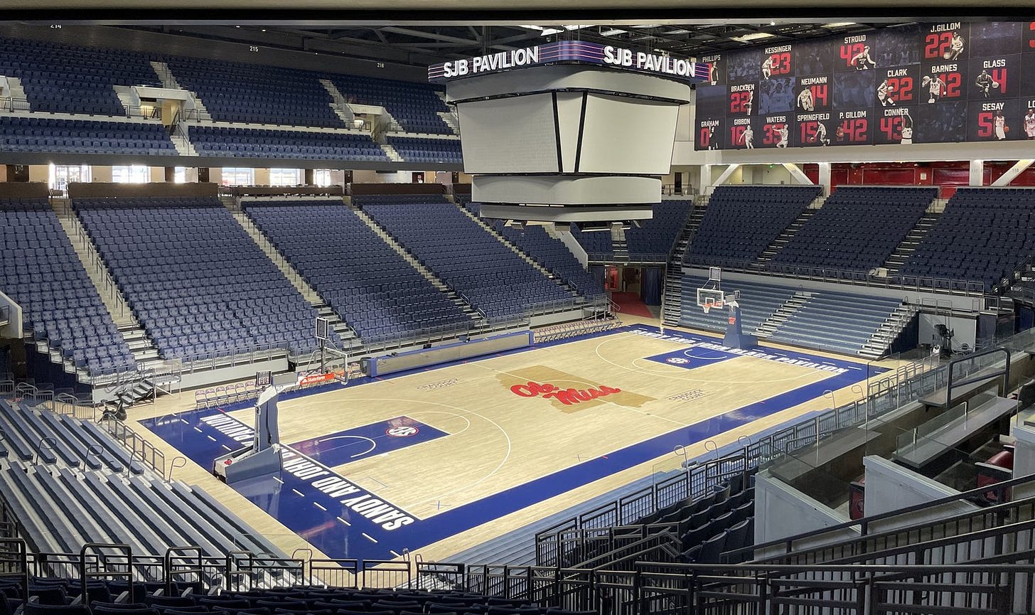 An empty indoor basketball arena seen from an upper level, with a wood court marked "Ole Miss" at center, blue seats on all sides and a scoreboard hanging under an "SJB Pavilion" sign. An empty indoor basketball arena seen from an upper level, with a wood court marked "Ole Miss" at center, blue seats on all sides and a scoreboard hanging under an "SJB Pavilion" sign.