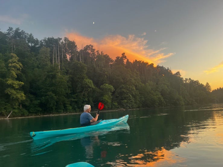A white-haired man with red paddles in a turquoise kayak on the Rhine river in evening light. Green trees grow along the riverbanks and the water reflect pink, sunset-colored clouds. A white-haired man with red paddles in a turquoise kayak on the Rhine river in evening light. Green trees grow along the riverbanks and the water reflect pink, sunset-colored clouds.