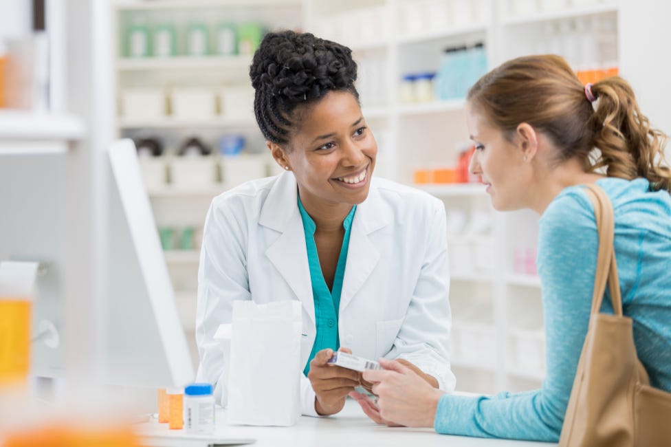 A Black woman pharmacist in a white coat talks with a woman customer at a pharmacy counter about her medication.