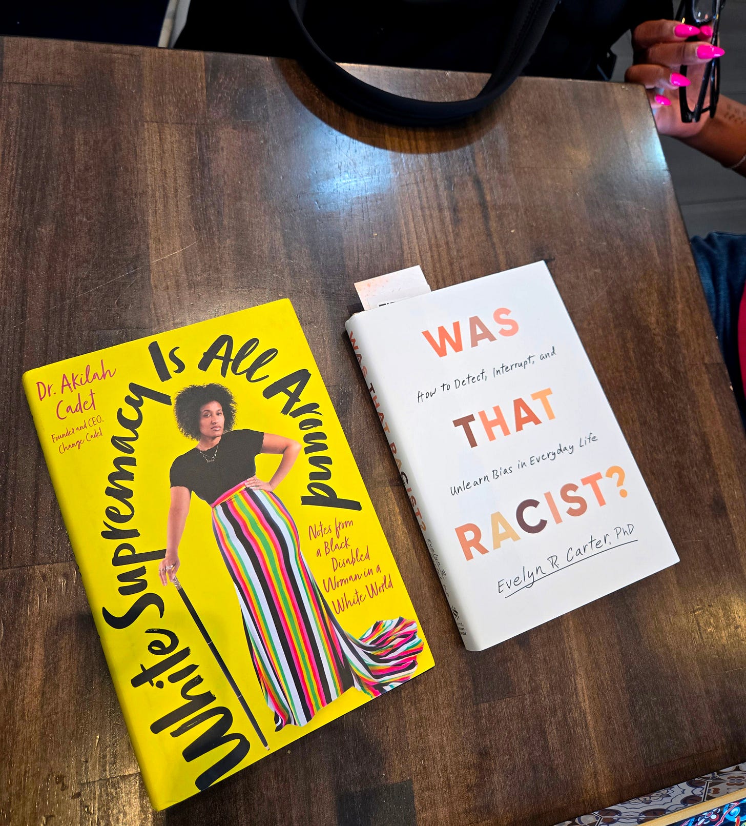 On a dark wood cafe table Dr. Cadet’s yellow book, White Supremacy Is All Around, and Dr. Carter’s white book, Was That Racist?, lay next to each other. You can see the perfect hot pink nails against Dr. Carter’s chocolate skin.