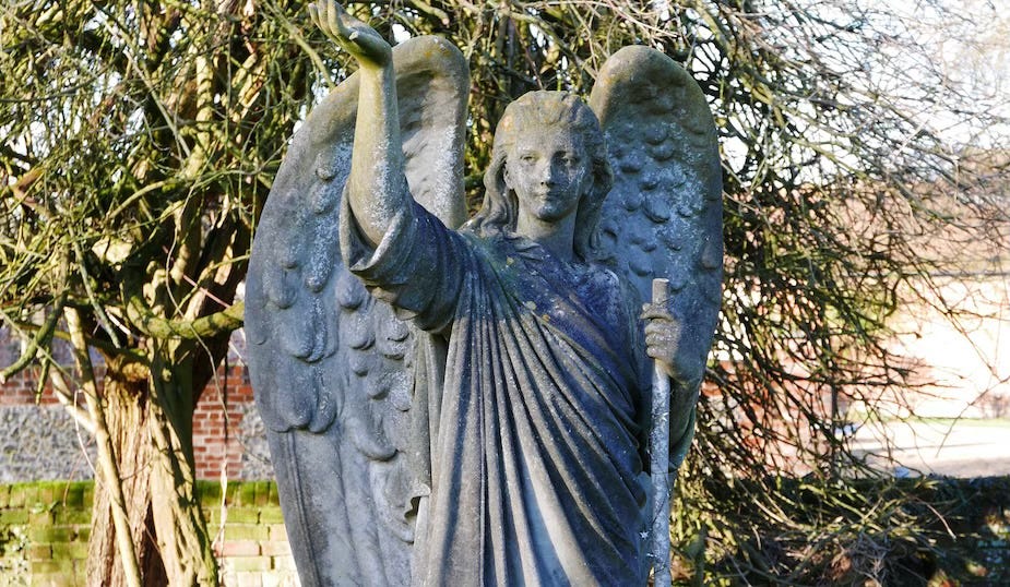 A weathered stone statue of an angel with large wings, standing outdoors in front of bare tree branches. The angel has long hair, a serene expression, and is dressed in draped robes. One arm is raised upwards, while the other holds a staff or column. Moss and age stains are visible on the surface, giving the figure an ancient, solemn appearance.