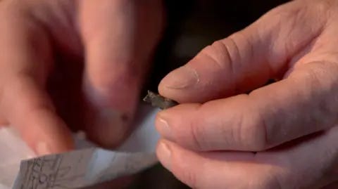 Kevin McGregor / BBC Ukrainian serviceman Serhiy Melnyk holds above a scrap of paper a small grey piece of shrapnel once lodged in his heart. Kevin McGregor / BBC Ukrainian serviceman Serhiy Melnyk holds above a scrap of paper a small grey piece of shrapnel once lodged in his heart.