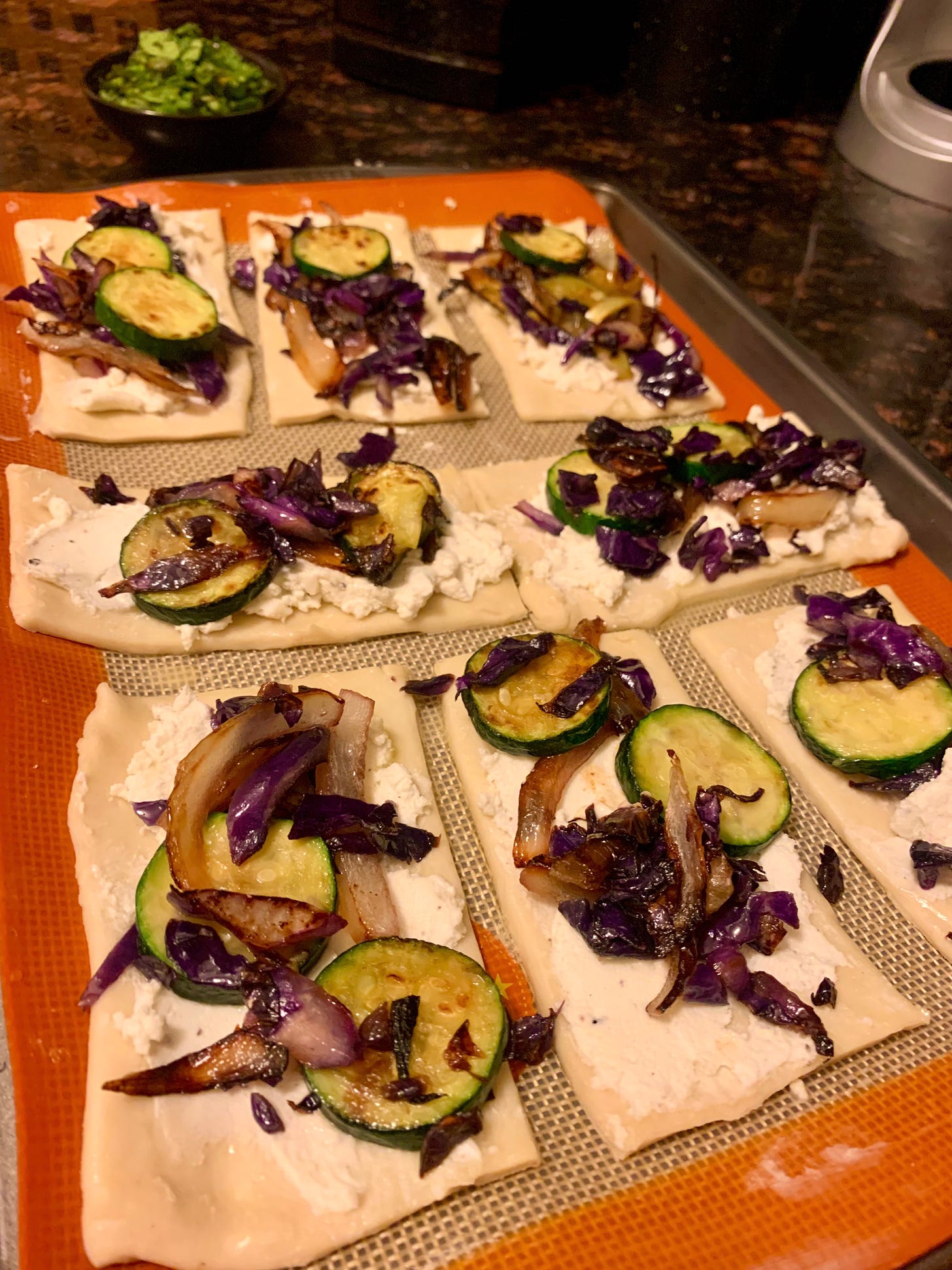 Angle view of puff pastry topped with vegetables and a small bowl of chopped mint in the background