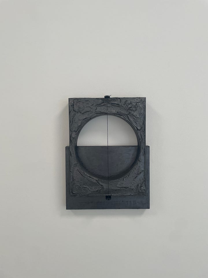 Gray, wooden sculpture carved into a rectangle with a large circle in the center, mounted vertically on a solid gray wooden panel that is mounted horizontally with a string cutting through the center of all of it vertically, all mounted on a white wall